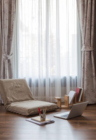 Big window of living room with sun light come through thin white curtain. Temporary area for online learning at home with foldable lazy chair, laptop, books and stationery on the floor.